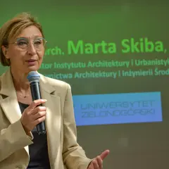 Kobieta w beżowej marynarce mówi do mikrofonu na tle zielonego ekranu z napisem "Uniwersytet Zielonogórski".
