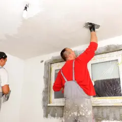 Dwóch budowlańców tynkuje sufit i ściany w pomieszczeniu.