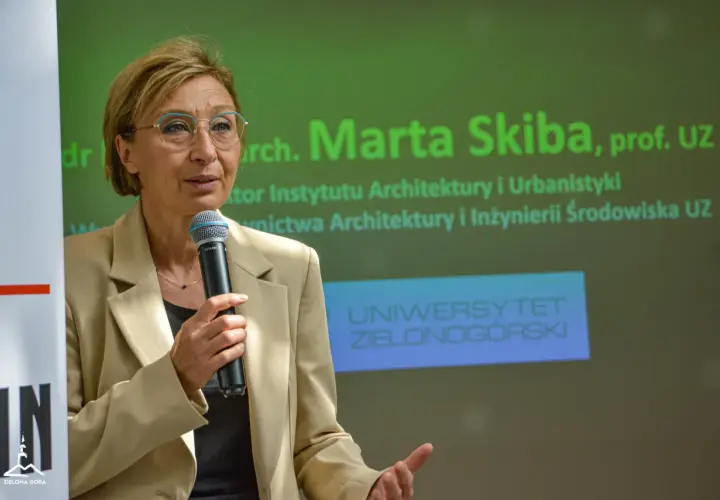 Kobieta w beżowej marynarce mówi do mikrofonu na tle zielonego ekranu z napisem "Uniwersytet Zielonogórski".