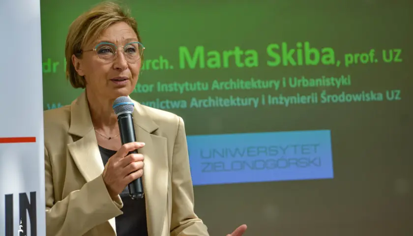 Kobieta w beżowej marynarce mówi do mikrofonu na tle zielonego ekranu z napisem "Uniwersytet Zielonogórski".